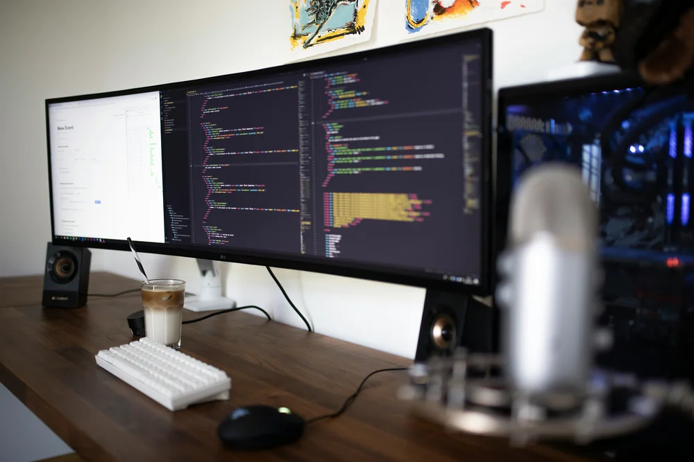 A wide-screen monitor, white keyboard and glass of iced coffee on a desk in an office