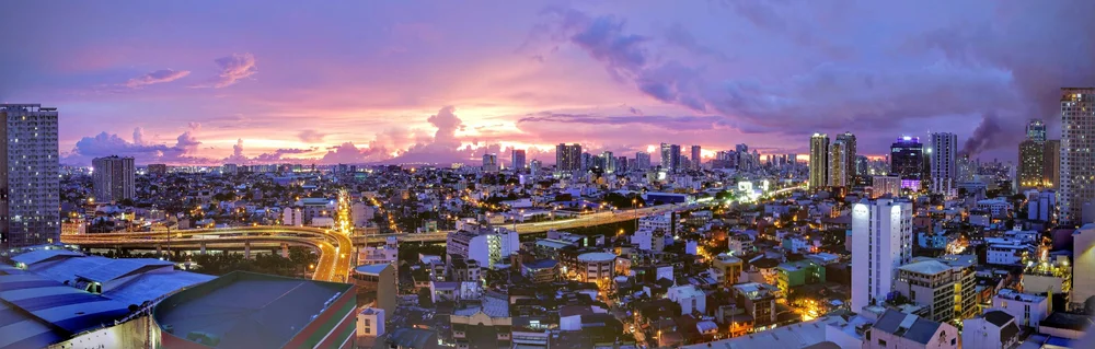 Wide panoramic cityscape of Manila, Philippines at sunset, with dramatic purple and pink clouds illuminating a densely built skyline. City streets, overpasses, and high-rise buildings stretch across the full frame, capturing the scale of the metro area.