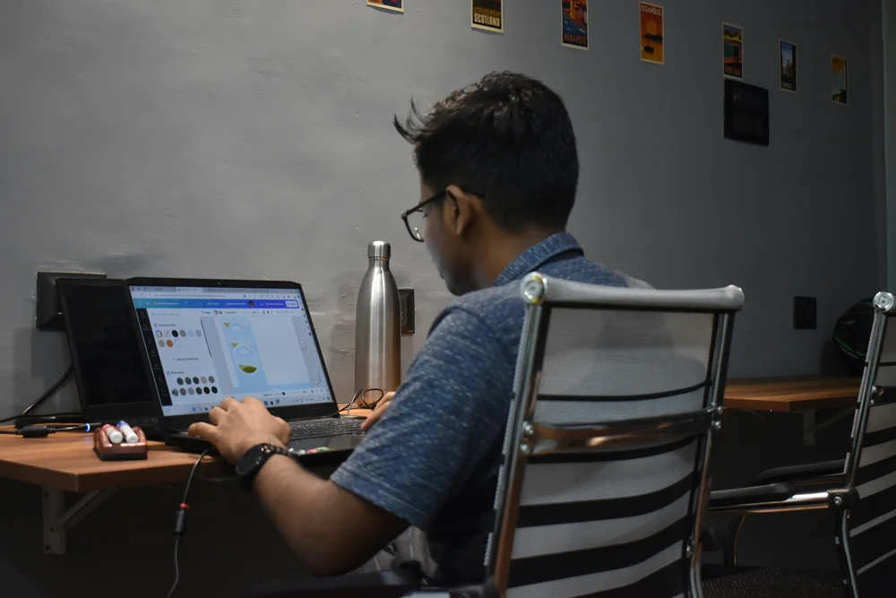 A young man wearing glasses works at a laptop in a dimly lit co-working space, viewed from behind. Travel posters from Scotland, Istanbul, and other destinations decorate the gray wall above him, and a metal water bottle sits on the desk beside the laptop.