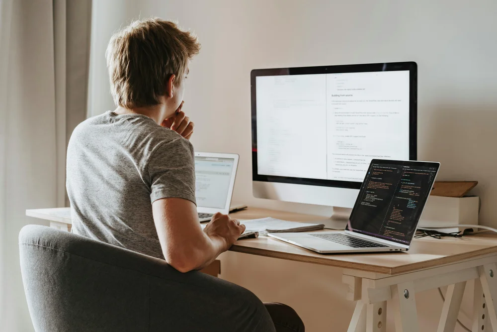A person in a grey t-shirt sits with their back to the camera at a home desk, studying a large monitor displaying a technical document alongside a laptop showing code, with a third laptop open to the left, depicting a multi-screen remote engineering setup.