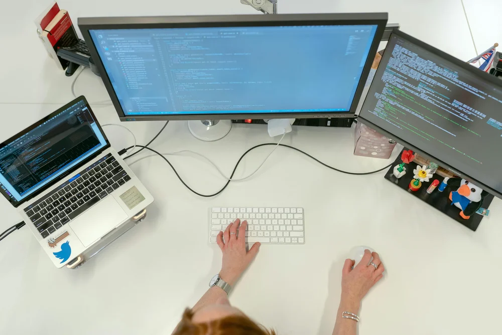 An overhead view of a person's hands typing on an Apple Magic Keyboard at a white desk, with a large monitor displaying code, a second monitor with terminal output, and a sticker-decorated laptop to the left, depicting a remote engineering workstation.