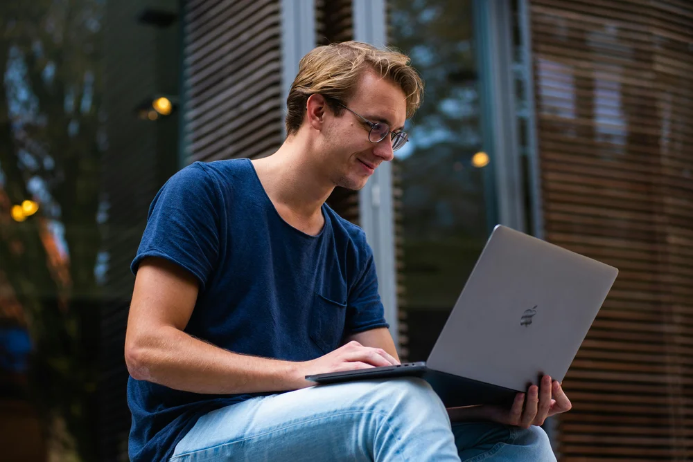 A young man wearing glasses and a blue t-shirt sits outdoors smiling while using a MacBook laptop, with a modern building facade and ambient string lights visible in the background, illustrating flexible remote work for engineers.