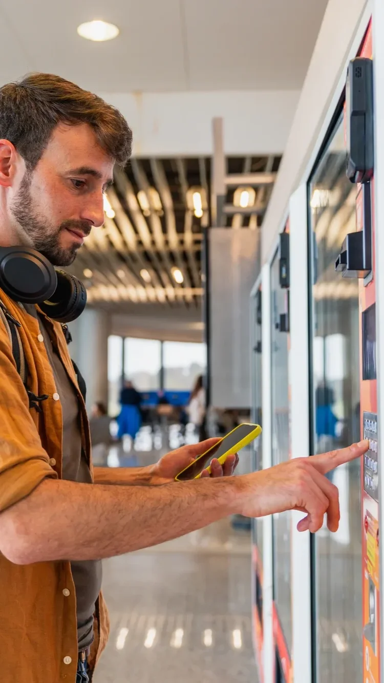 Mann mit Kopfhörern und Smartphone bedient den Touchscreen eines Verkaufsautomaten in einer Halle