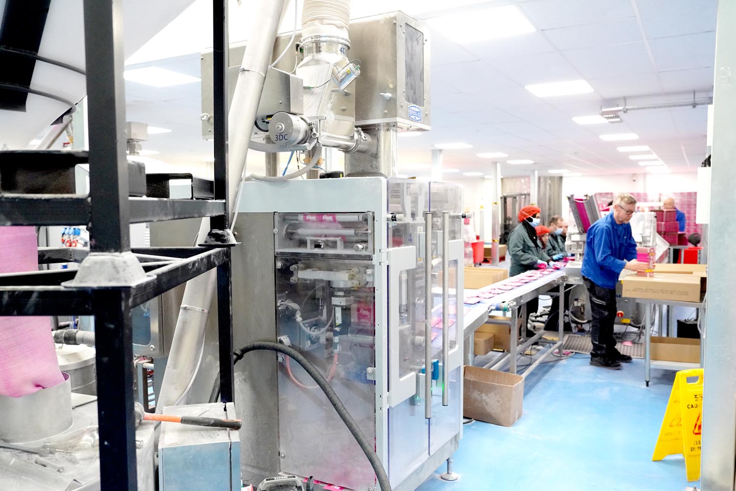 Workers in a factory packaging pink products on a conveyor belt, with machinery and a caution wet floor sign in the foreground.