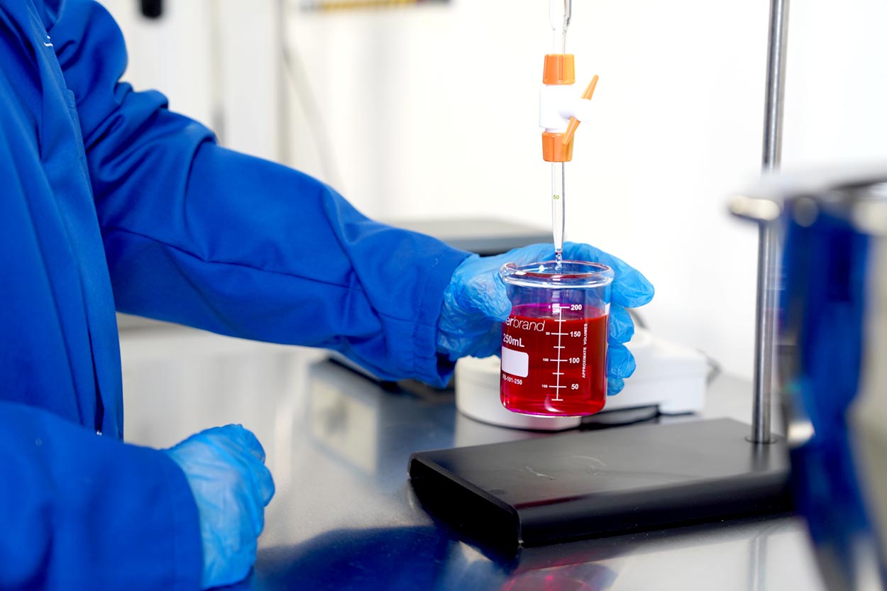 Scientist wearing blue gloves and lab coat holding a beaker with red liquid while dispensing a dropper.