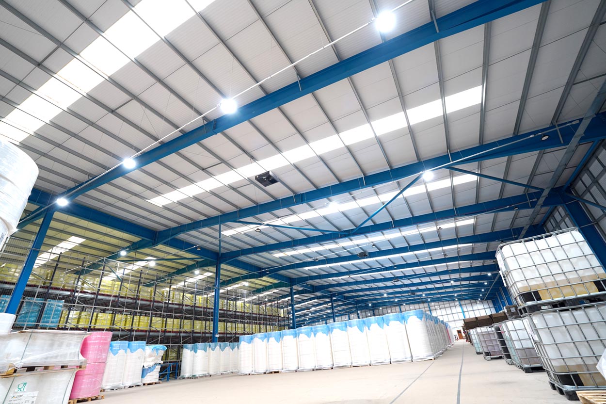 Large industrial warehouse interior with blue steel beams, bright lights, and neatly stacked white and colored pallets and containers.