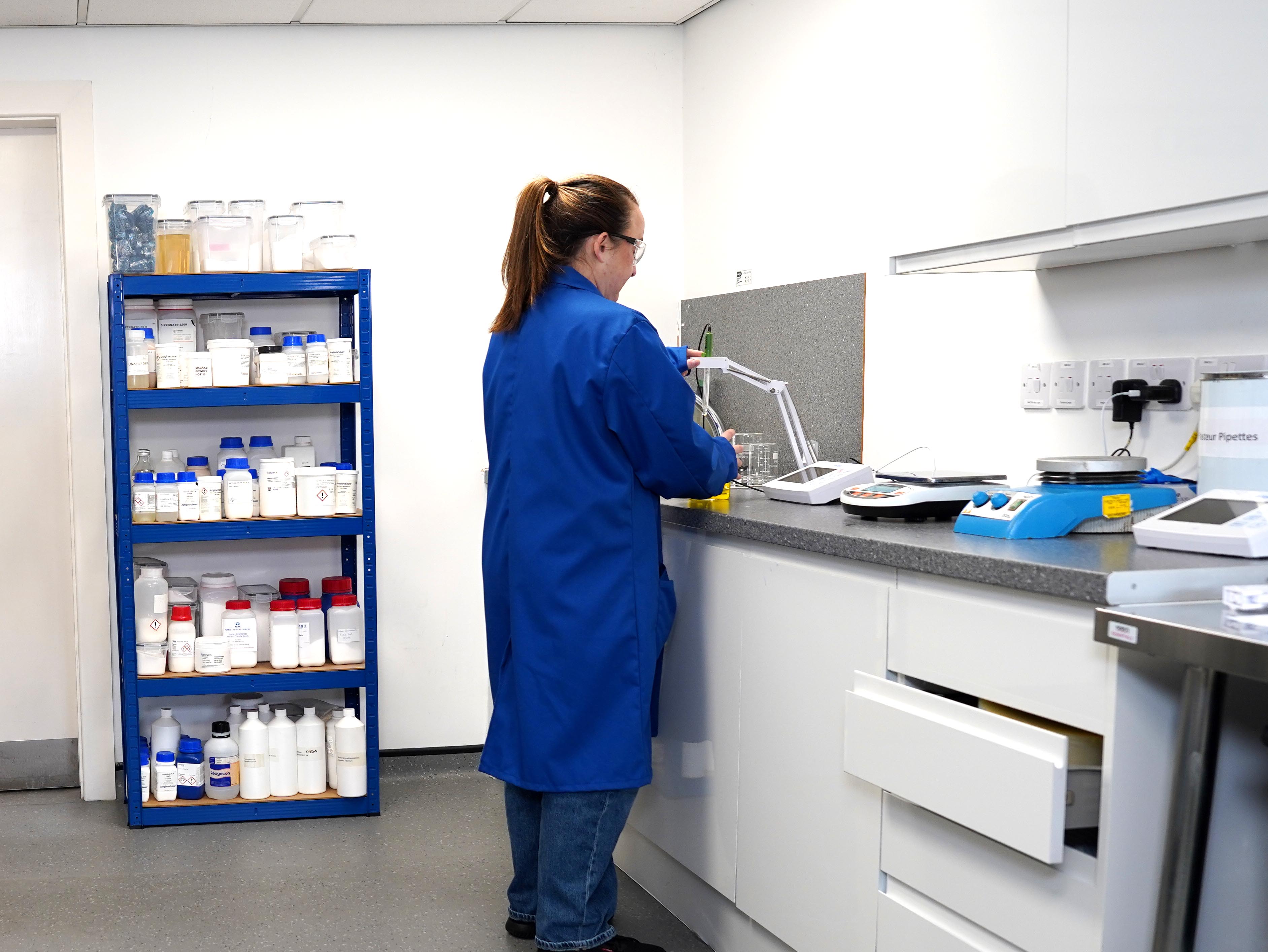 Scientist in blue lab coat working with glassware and equipment on a laboratory counter, with shelves stocked with chemical containers in the background.