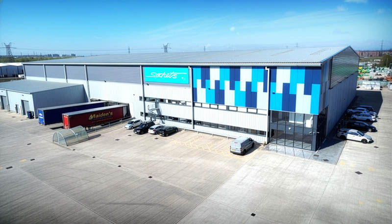 Large industrial warehouse with blue and white facade, parked cars, and a red Maiden's trailer truck outside under clear sky.