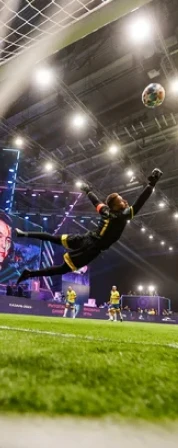 Soccer goalkeeper in black and yellow diving to save a ball near the goal on an indoor field.
