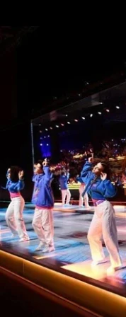 Four female performers in blue tops and pink pants dancing on a stage with colorful lighting and an indoor audience in the background.