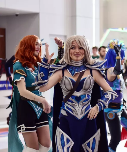 Two women dressed in elaborate fantasy cosplay costumes at a convention, one with red hair and green and black outfit, the other with blonde hair in silver and blue armor.