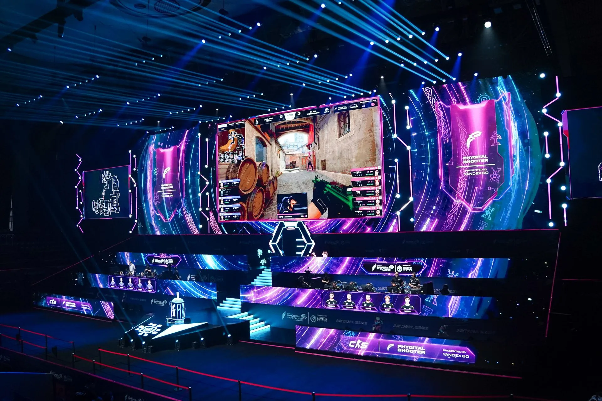 Large indoor arena filled with a crowd watching a brightly lit esports tournament on elevated central stage with multiple screens.