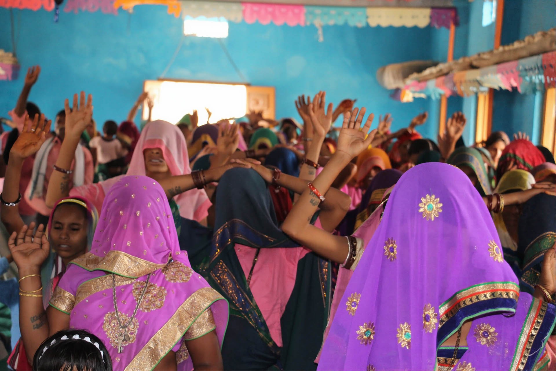 Group of women dressed in colorful sarees with covered heads raising their hands in a blue-painted room.