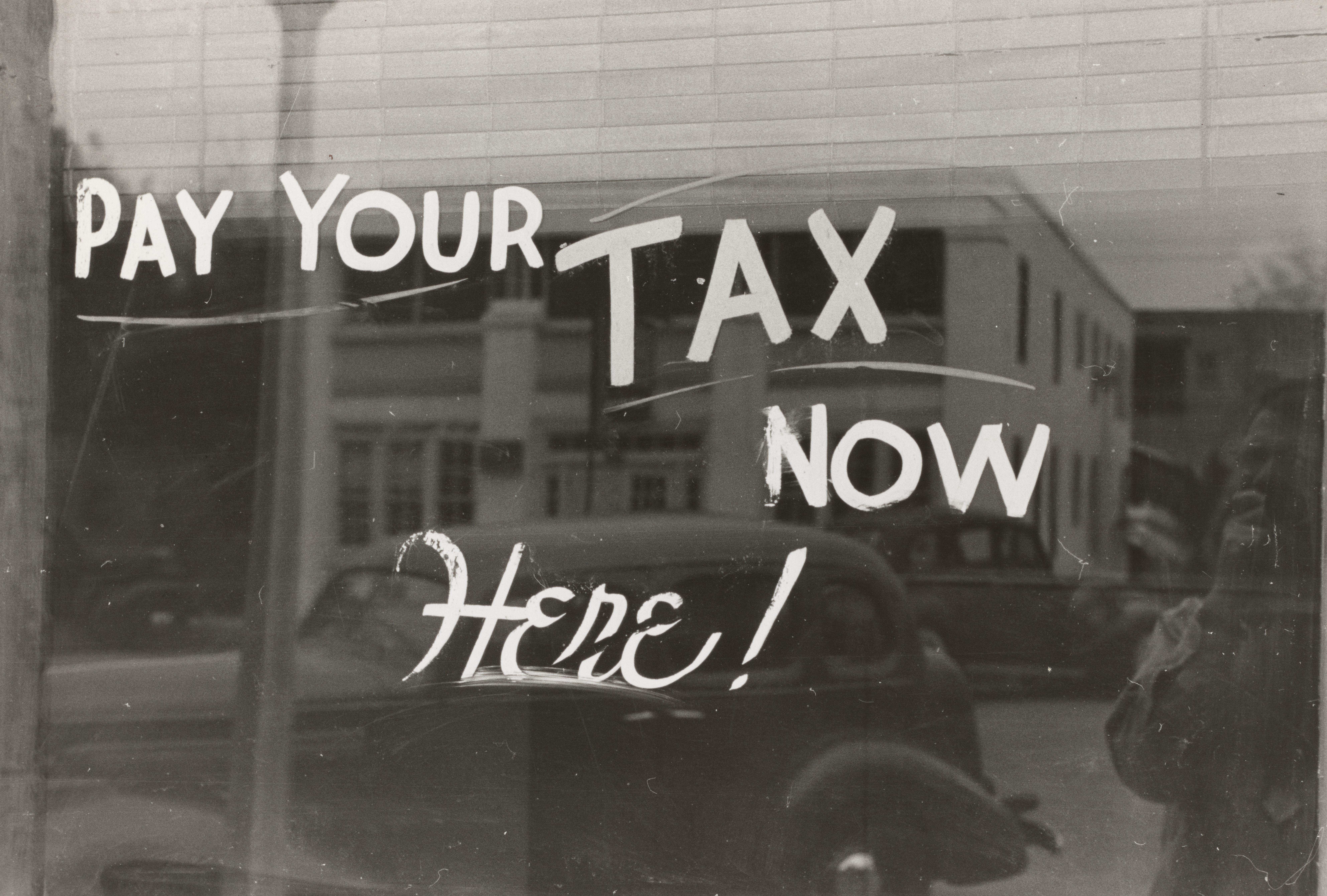 An antique display in sepia with a note on paying your taxes here now, painted on the window of what appears to be a storefront, with the bustle of Main Street reflecting on the mirrored background of the sign.