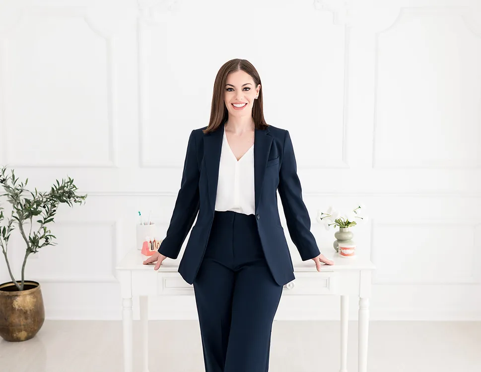 Smiling woman in navy blue suit and white blouse leaning against a white desk in a bright room with a potted plant and flowers.