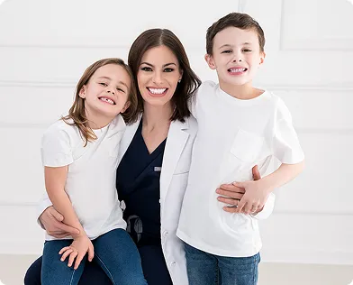 Smiling woman in a white coat with two children, one on each side, all wearing casual clothes and posing indoors against a white background.