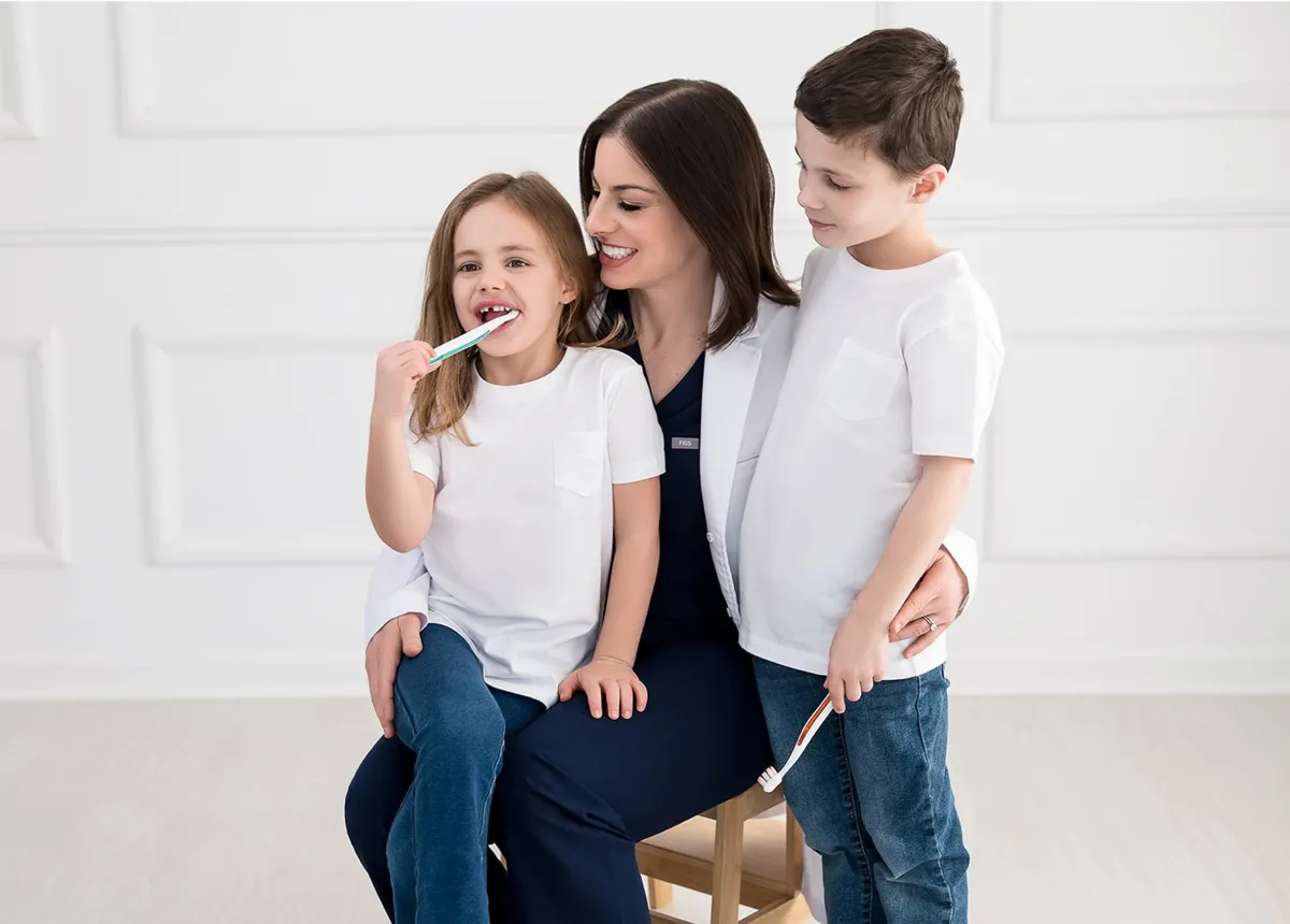 Female dentist sitting on a stool with a girl brushing her teeth and a boy holding a toothbrush.