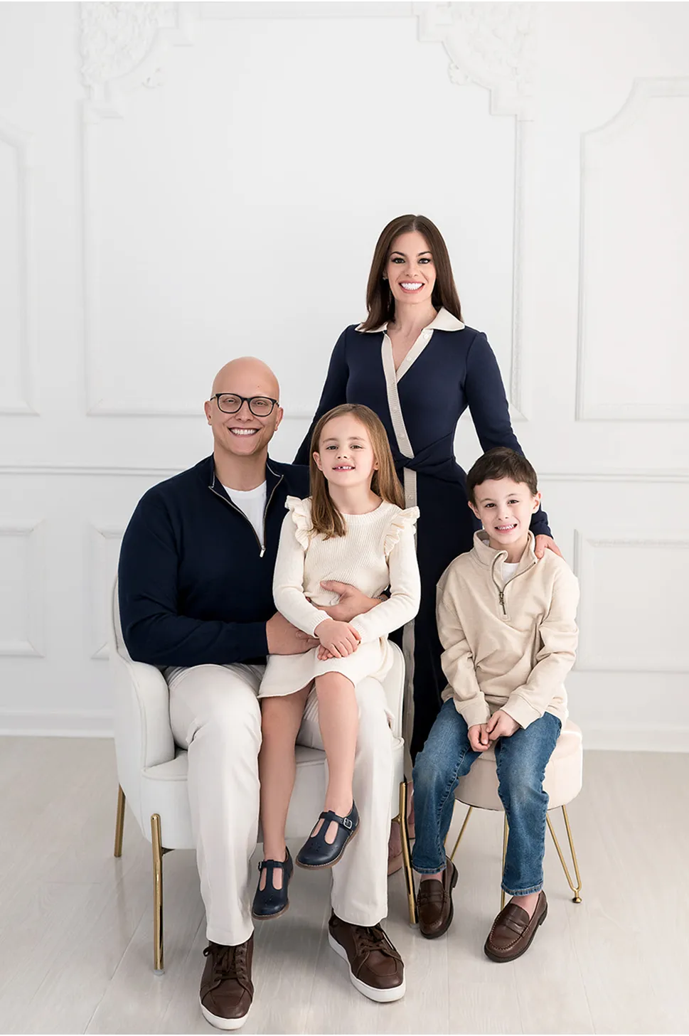 Smiling family of four with father sitting holding daughter, mother standing behind them, and son sitting on a stool against a white decorative wall.