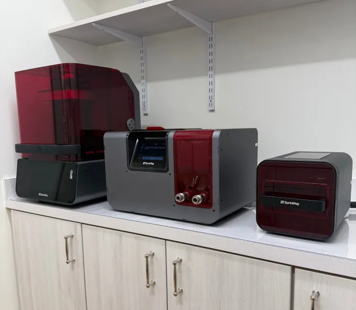 Three SprintRay 3D printers with red translucent covers placed on a countertop beneath a white shelf.