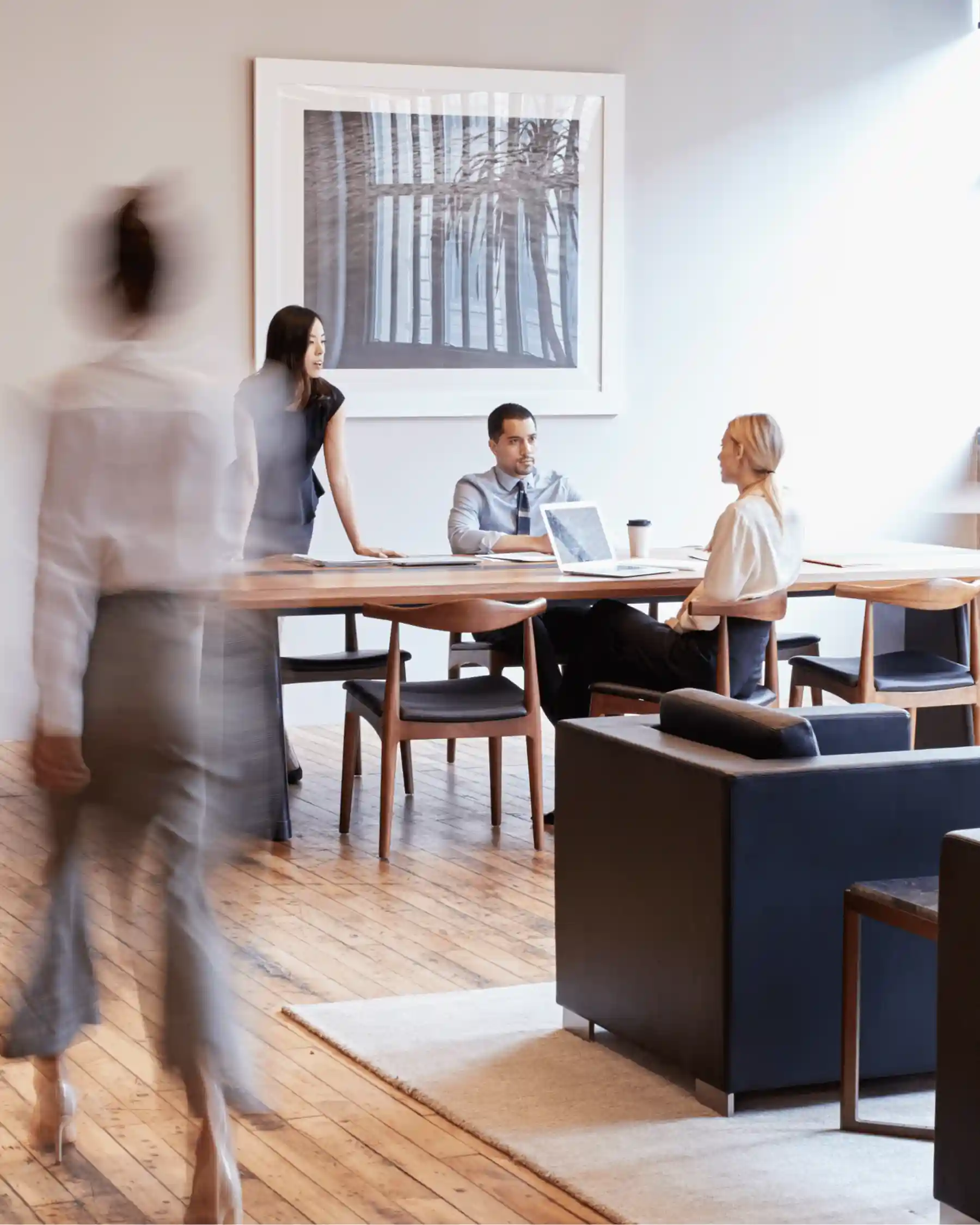 Three business people having a meeting at a wooden table in a modern office with a framed abstract artwork on the wall.