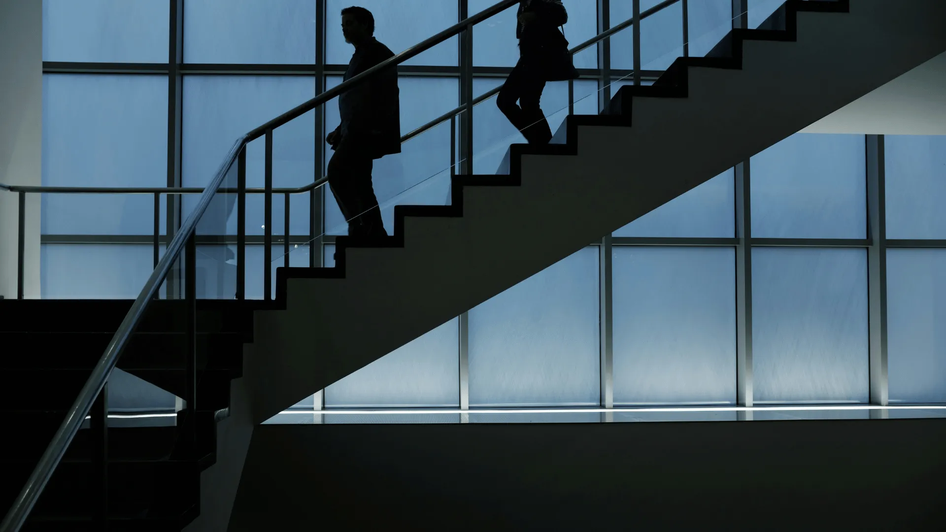 Silhouettes of two people walking down an indoor staircase with large windows in the background.