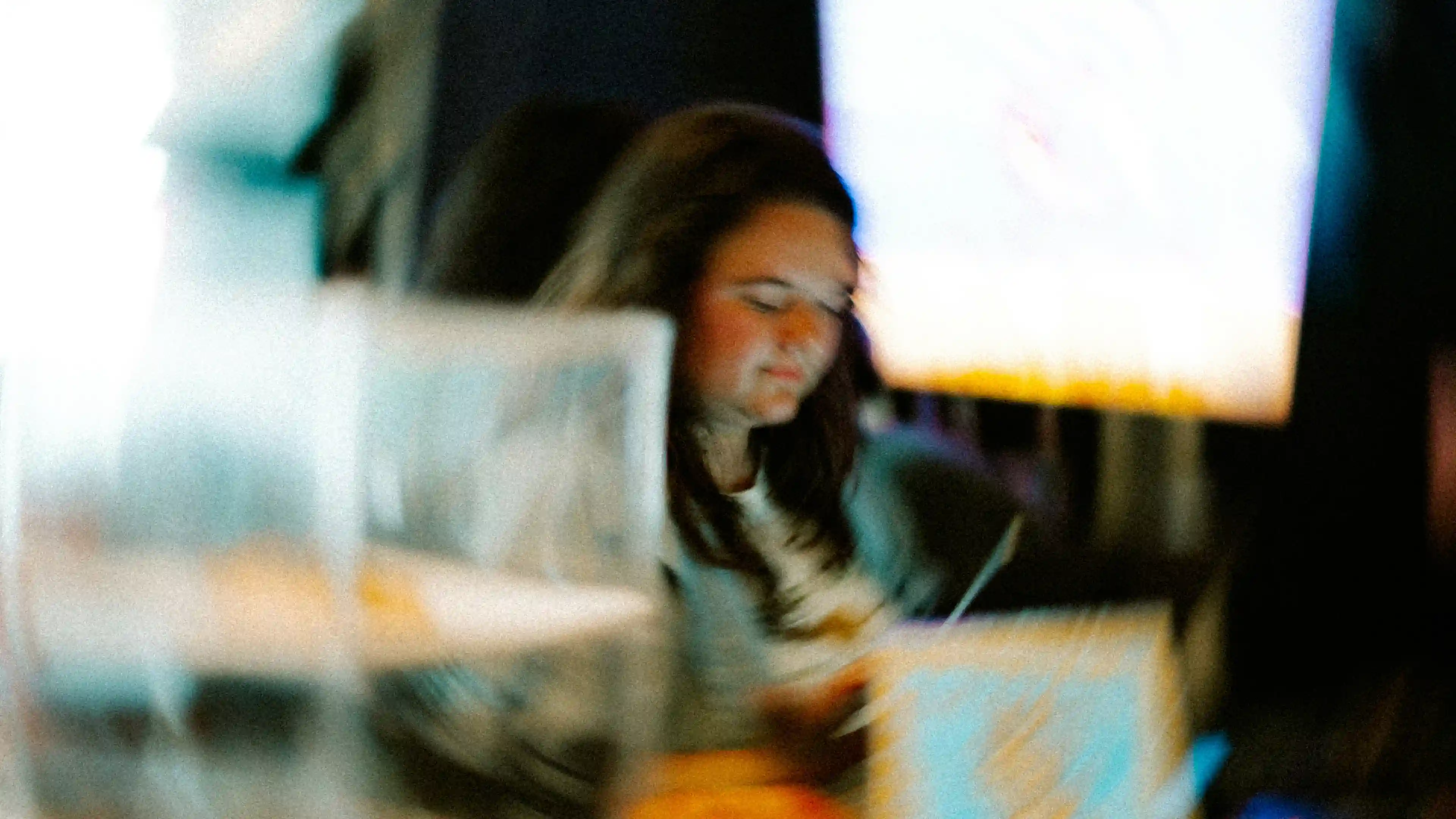 Blurred image of a person with long hair reading or writing, with bright screens or lights in the background.