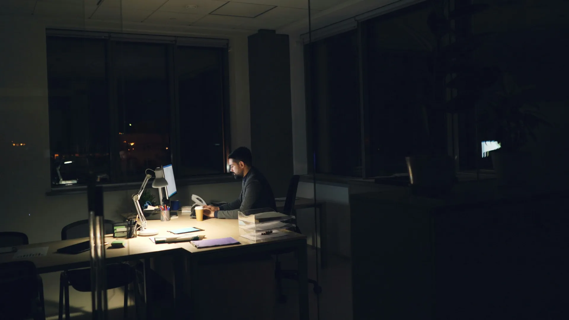 Man working late at a desk in a dark office illuminated by a desk lamp and computer screen.