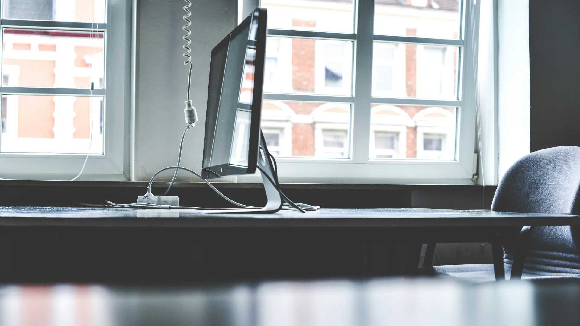 Side view of a computer monitor on a desk near a window with a chair nearby.