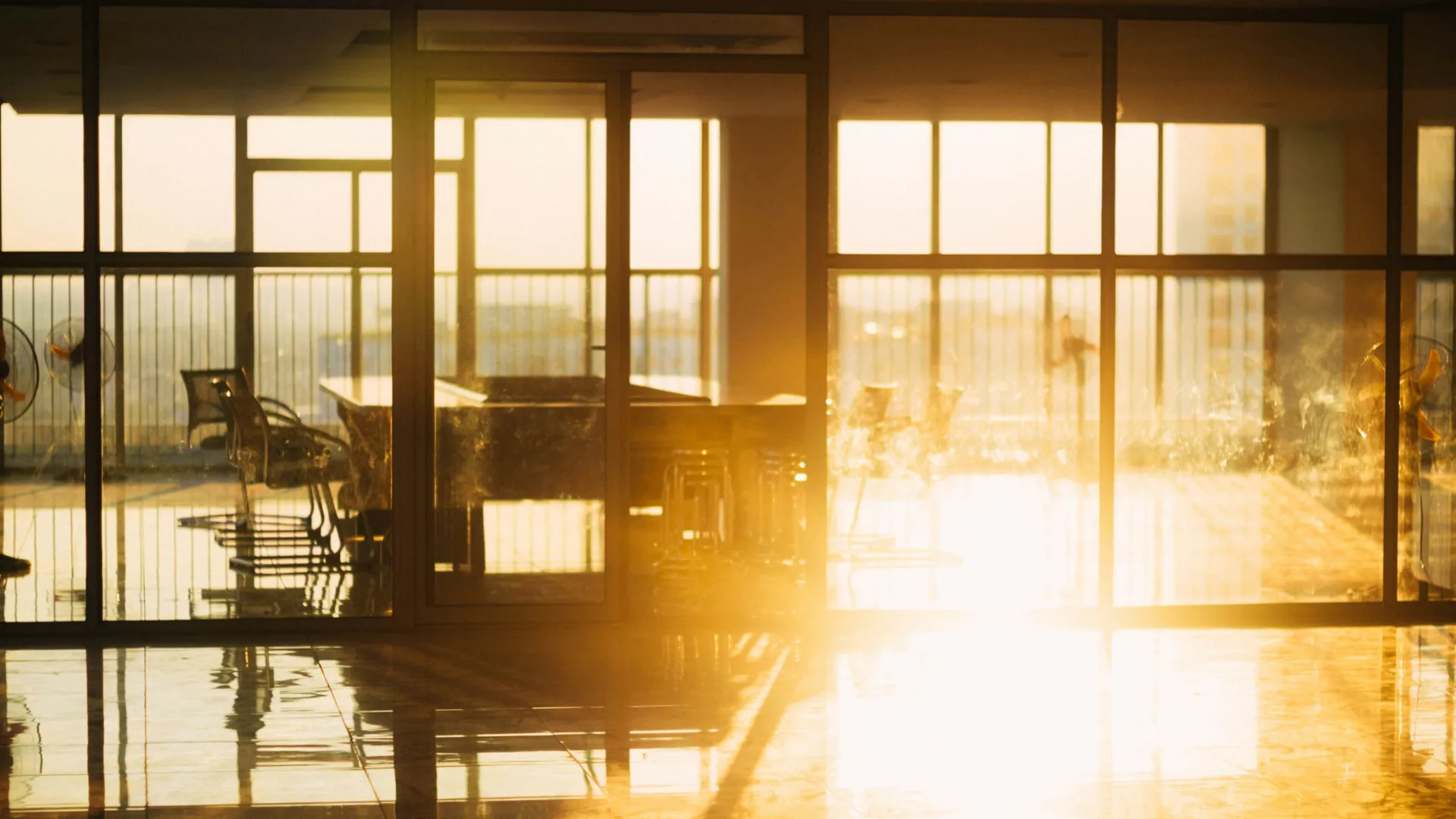 Sunlight flooding through large glass windows onto a modern office space with chairs, tables, and standing fans.