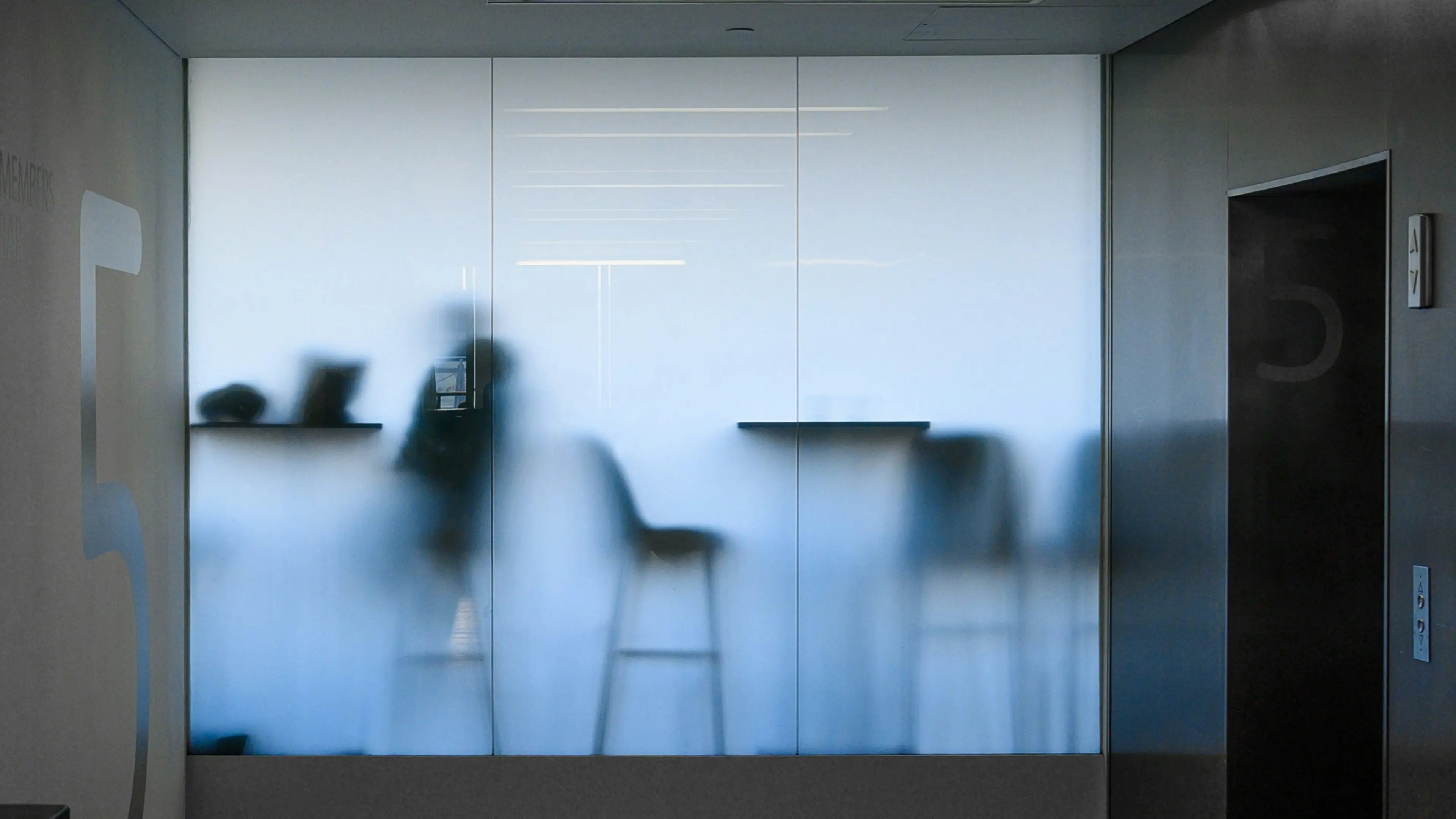 Blurred silhouette of a person sitting on a bar stool behind frosted glass in a modern room with the number 5 on walls.