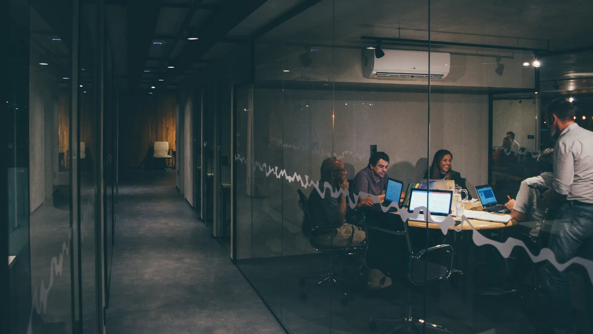 Group of colleagues having an evening meeting with laptops around a round conference table in a glass-walled office.