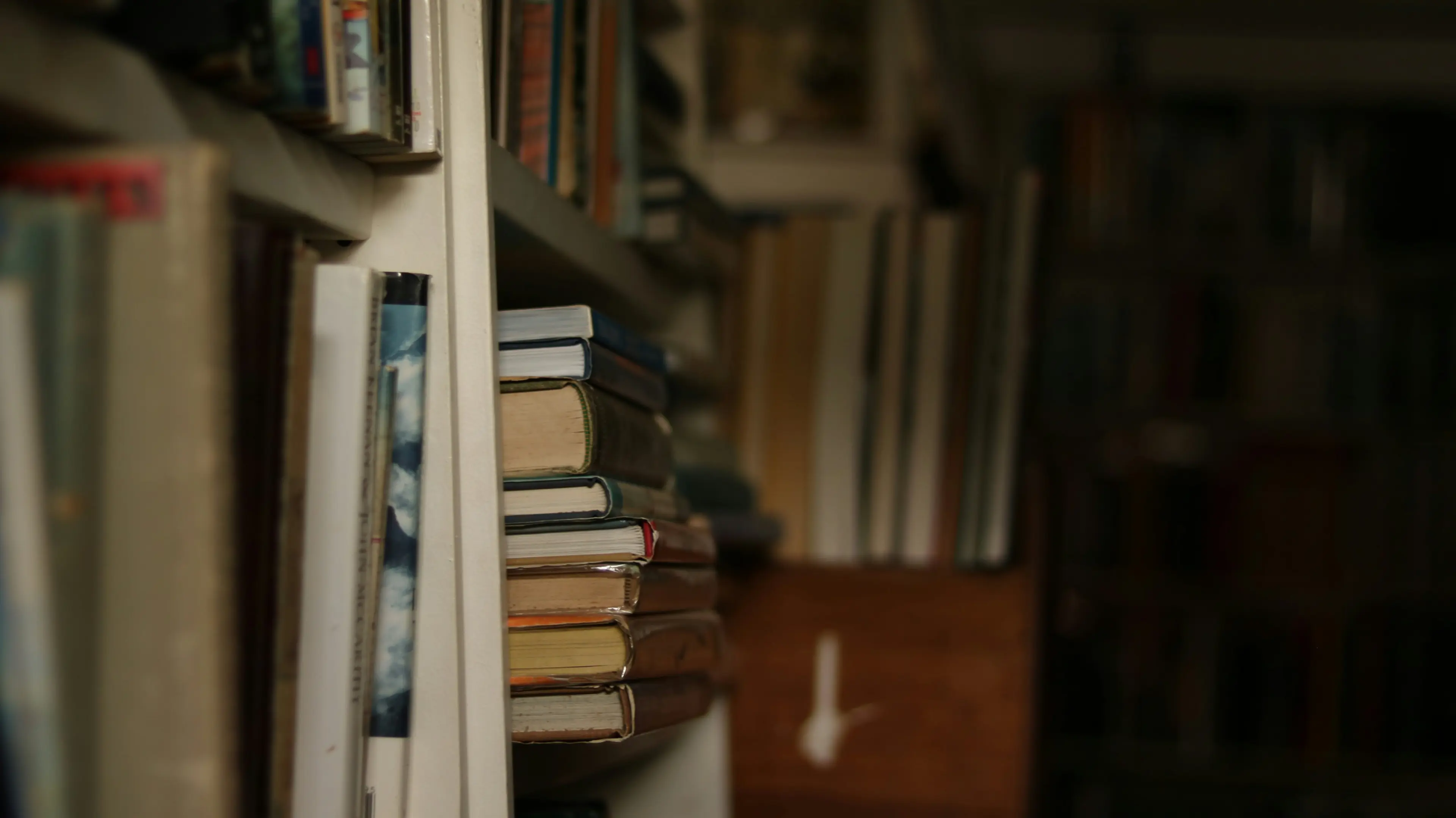 Side view of a stack of various hardcover books placed horizontally on a shelf with other books around them.