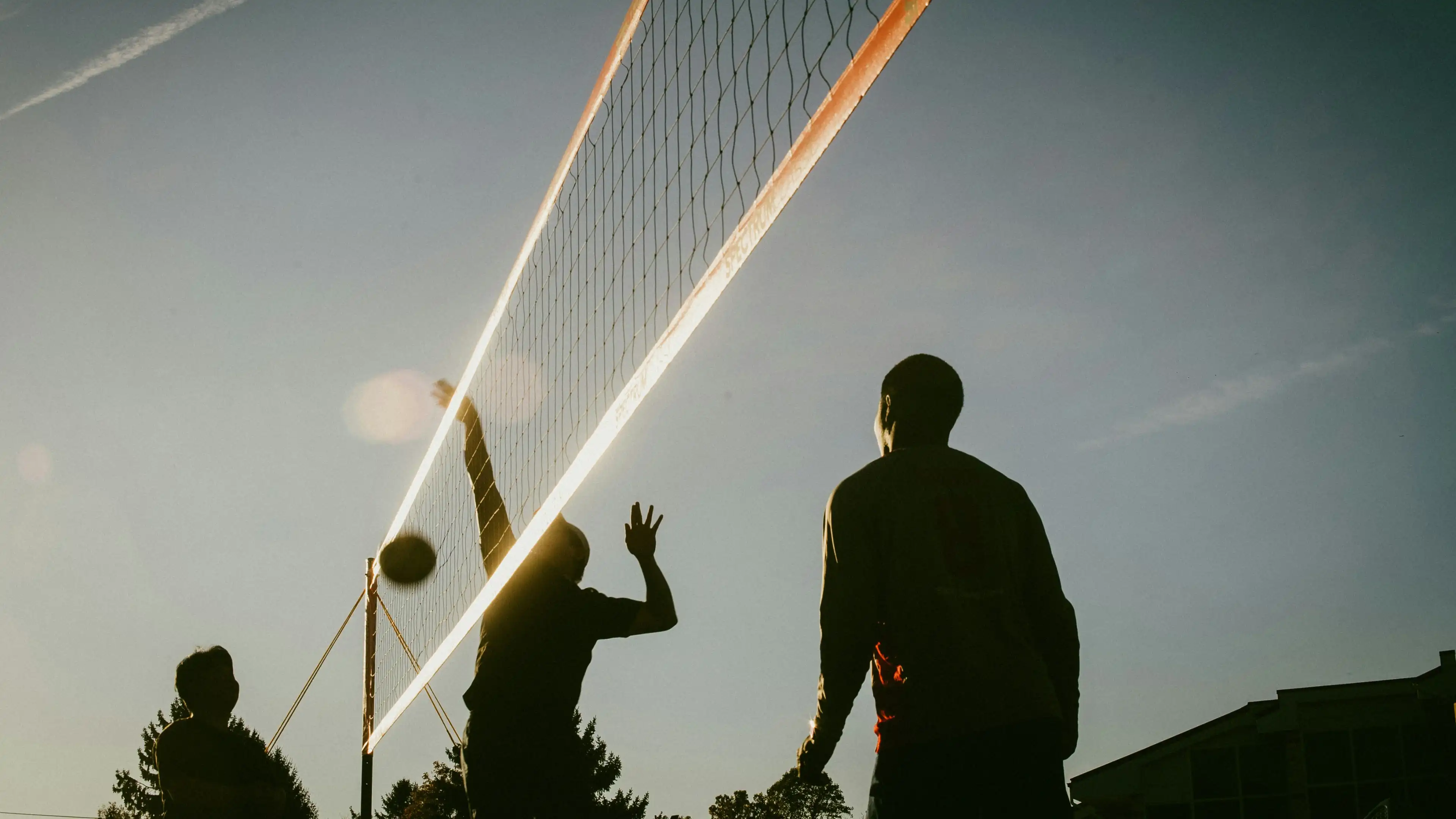 Silhouettes of three people playing volleyball at sunset with the ball near the net.