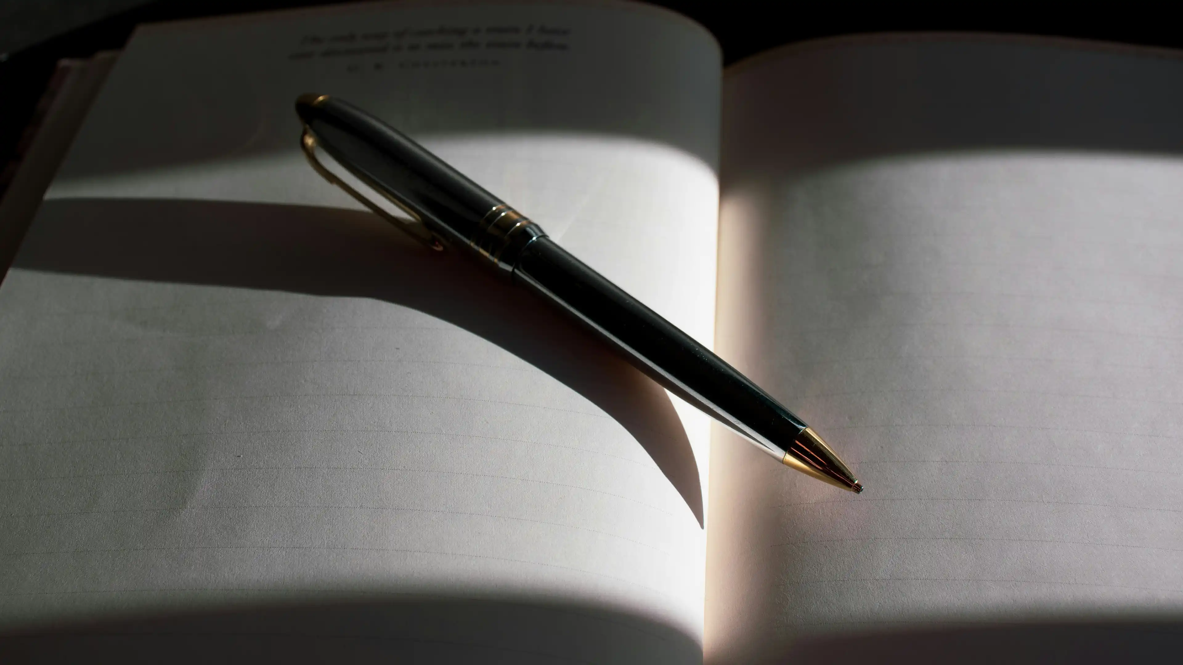 Black and gold pen resting diagonally on an open blank notebook with lined pages in soft lighting.