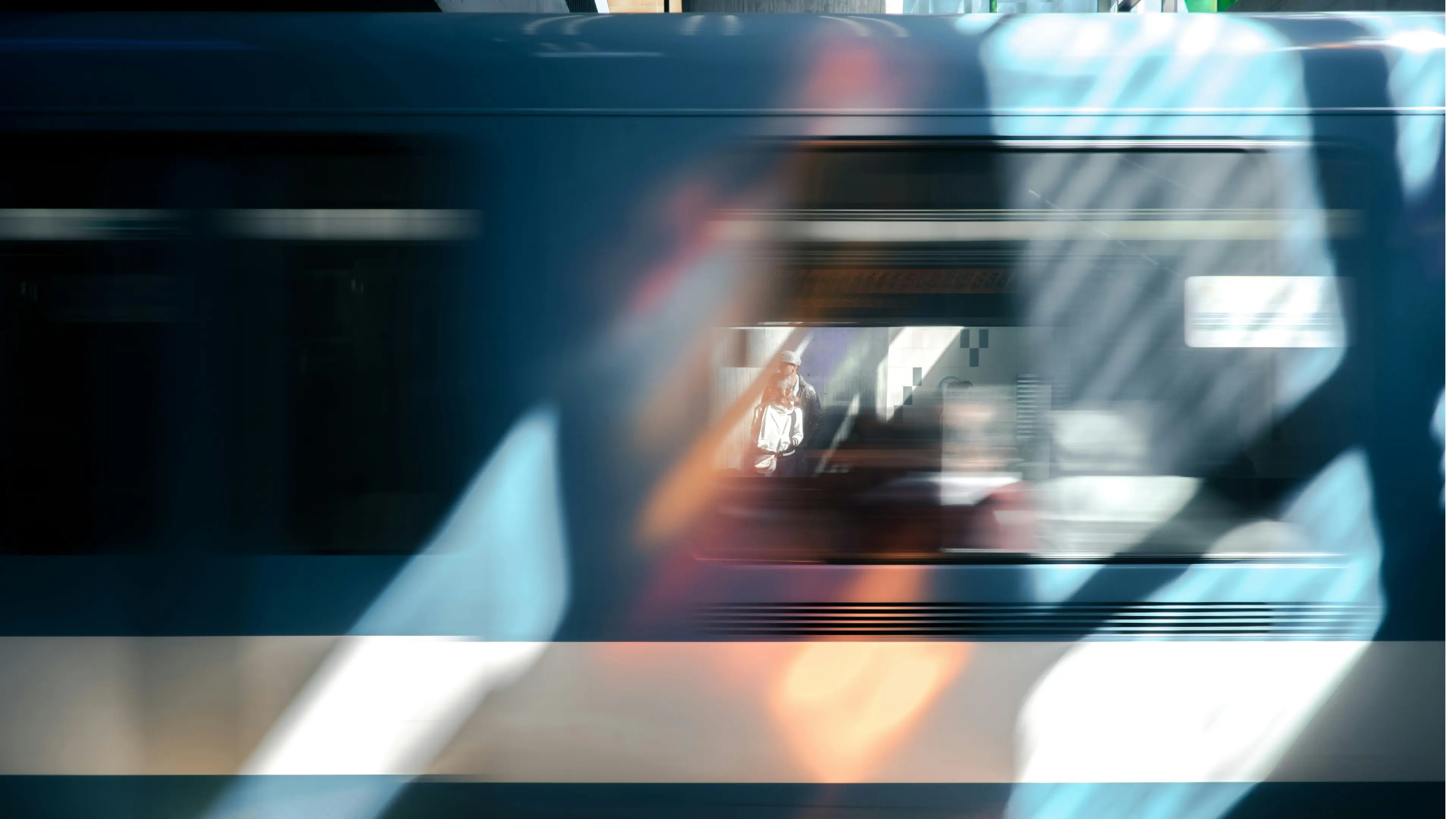 Blurred side view of a moving blue train with two people visible inside the station behind it.