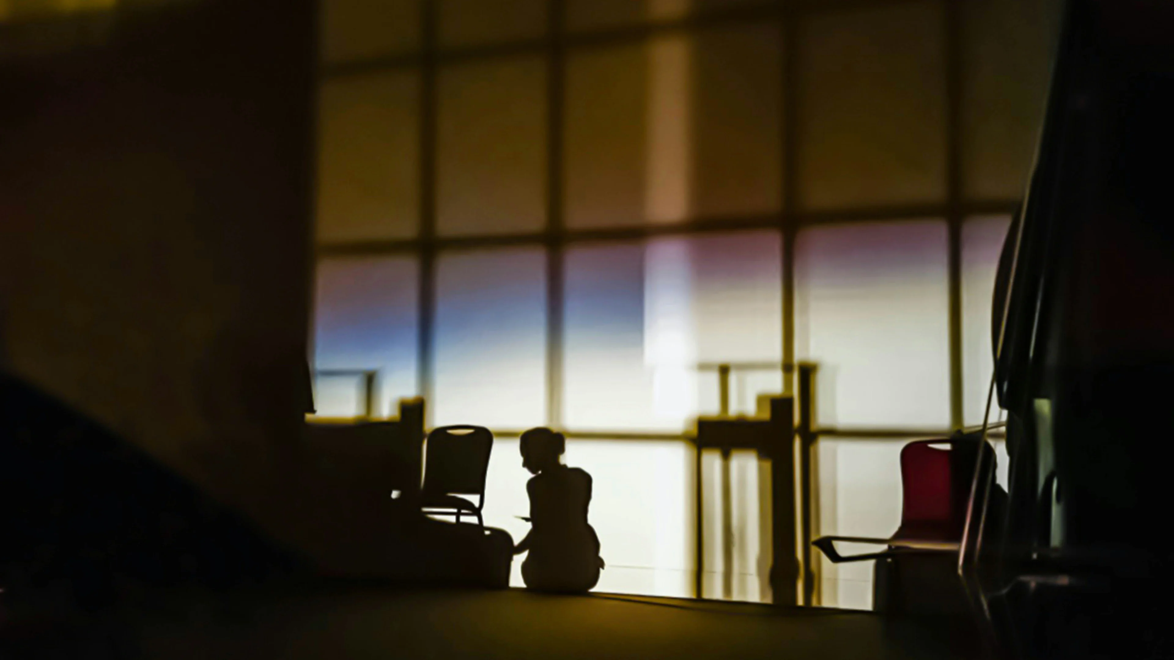 Silhouette of a person sitting on the floor inside a room with large grid windows, along with several chairs and furniture shadows.