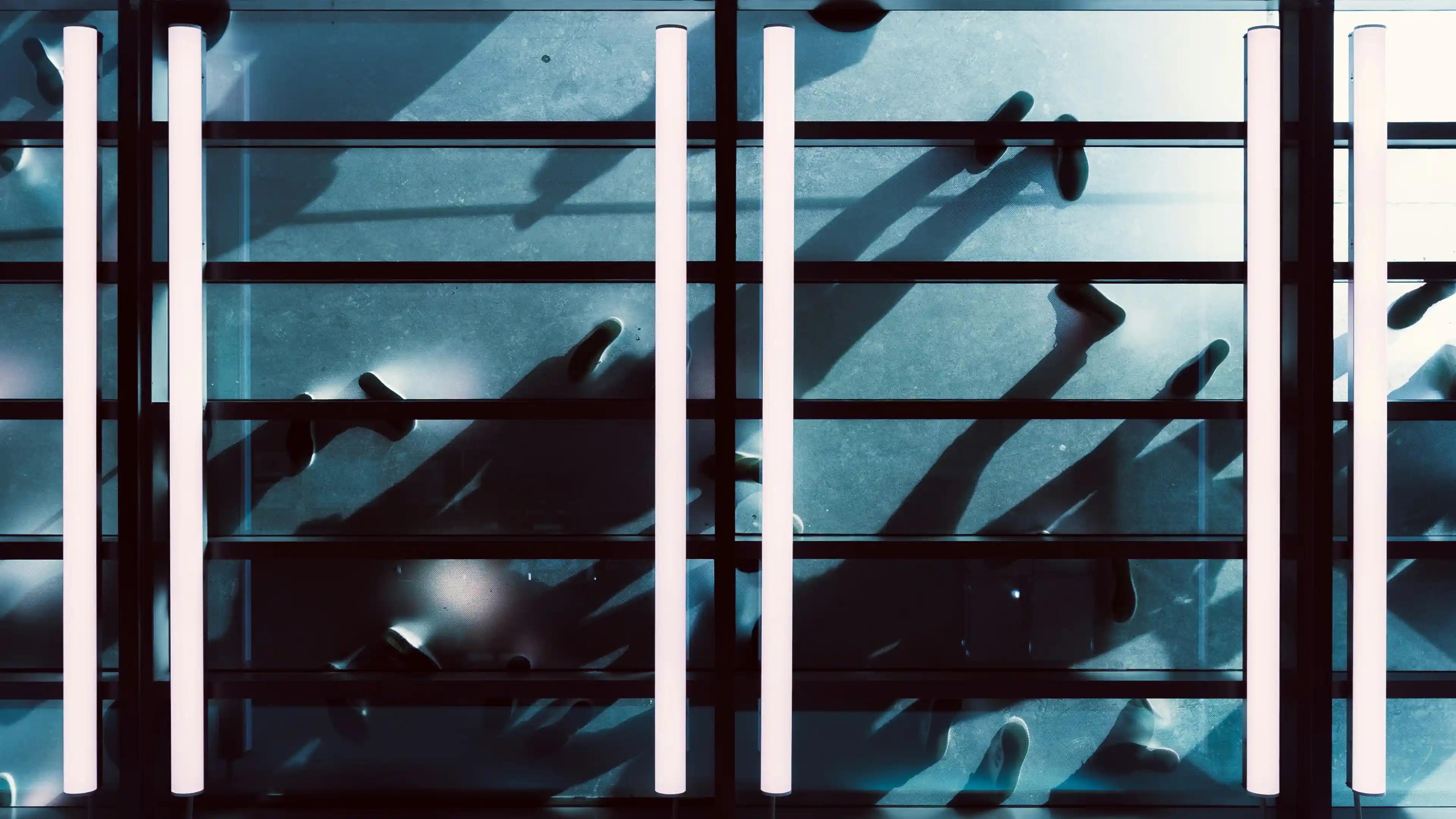 Overhead view of multiple people walking on a glass floor with strong shadows and white vertical light fixtures.