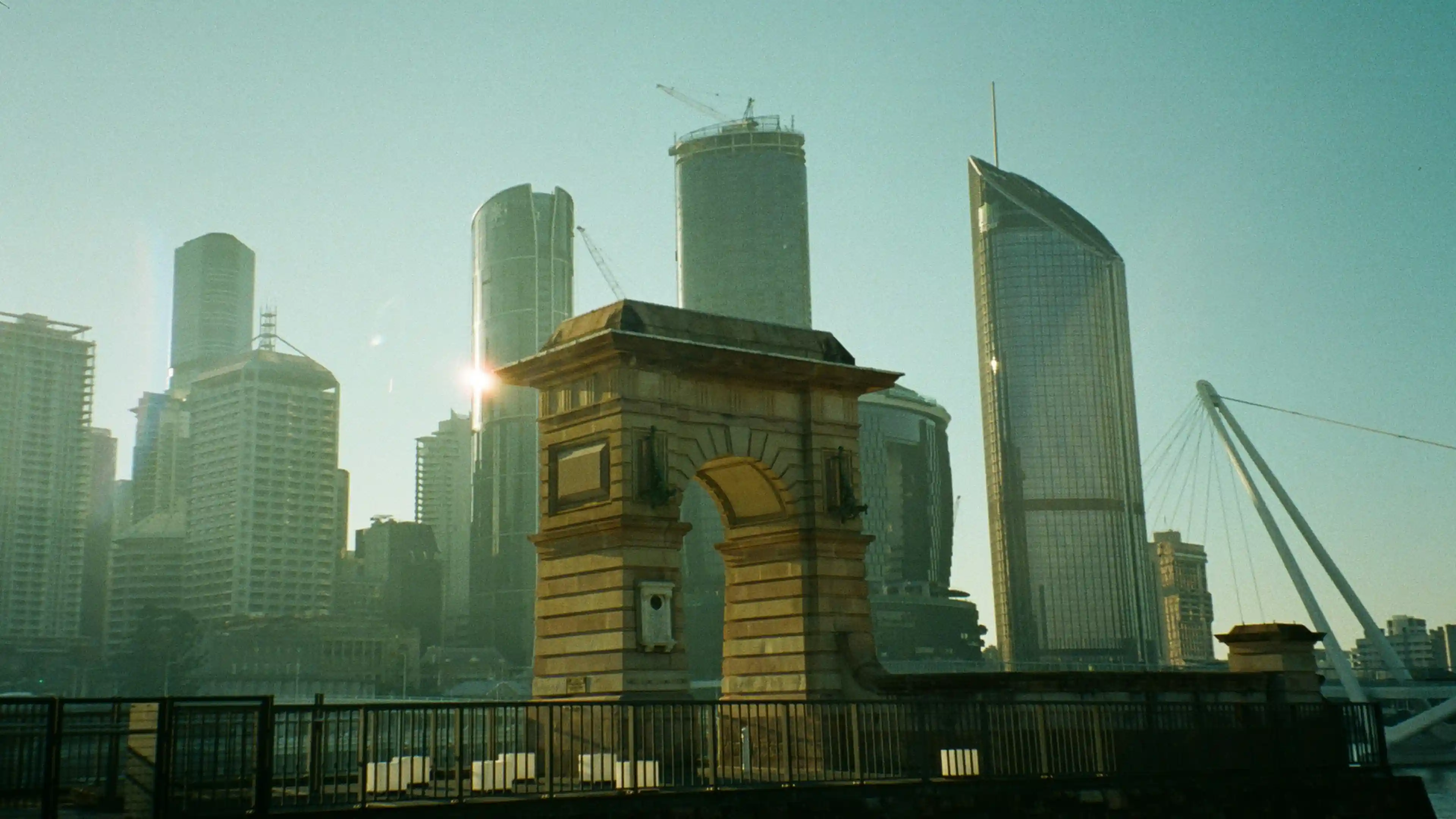 Stone arch monument with modern high-rise buildings and a suspension bridge in the background during daylight.