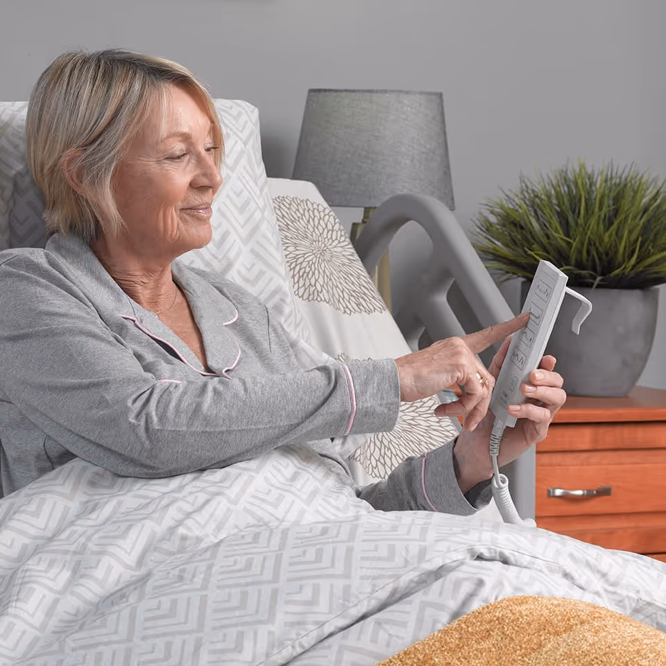 Elderly woman in gray pajamas sitting in a hospital bed adjusting a bed control remote.