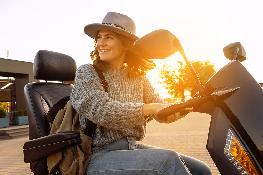 Smiling woman in a hat and sweater sitting on a mobility scooter outdoors at sunset.