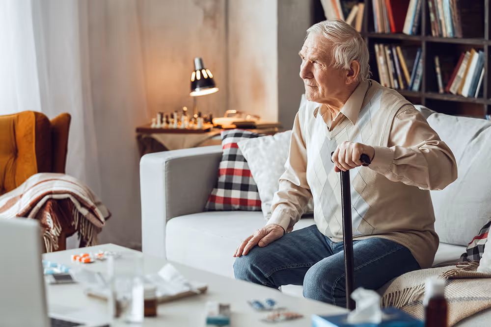 Elderly man holding a cane sitting on a couch in a cozy living room with books and chess set.