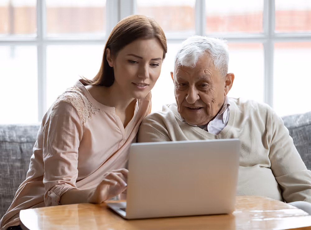 Young woman helping elderly man use a laptop at a table near a window.
