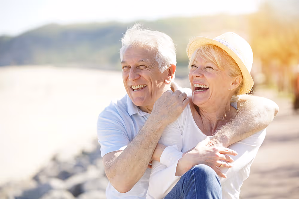 Smiling elderly couple hugging outdoors on a sunny day near a beach path.