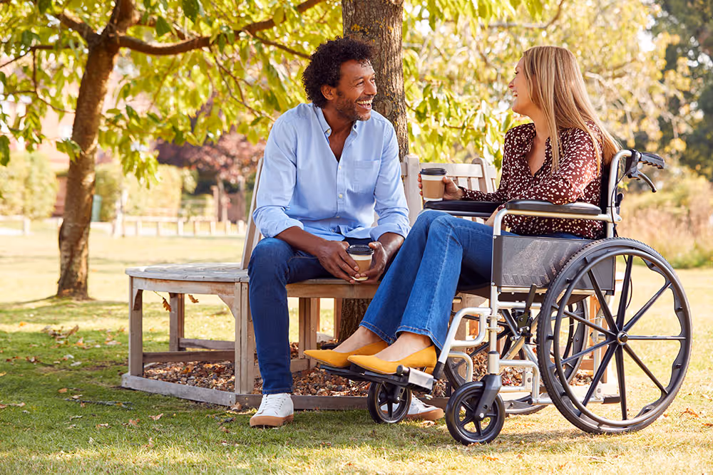 Man and woman in a wheelchair enjoying coffee and chatting outdoors under a tree on a sunny day.