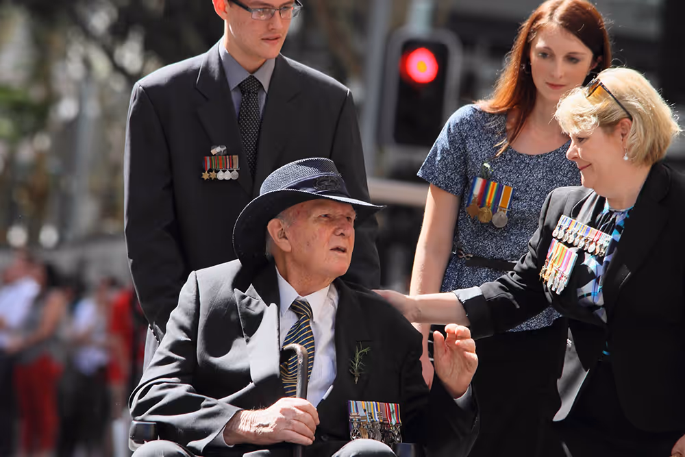 Elderly veteran in wheelchair wearing a hat and medals, accompanied by two women and a man with medals.