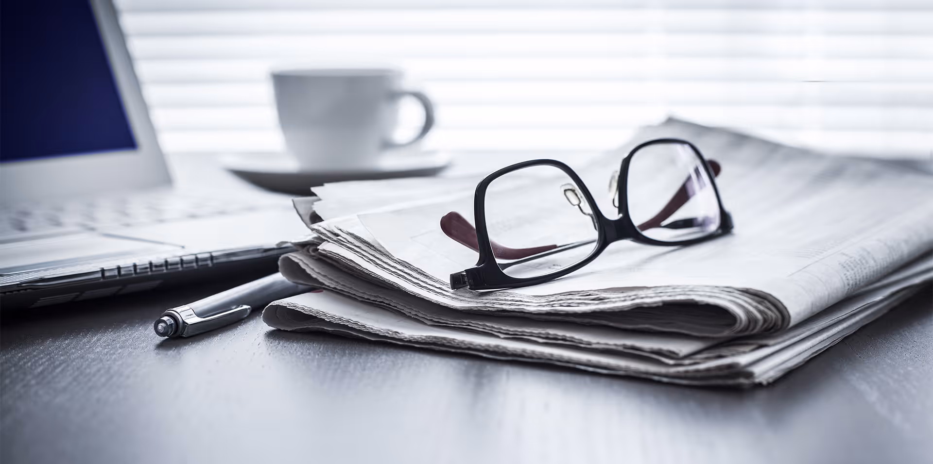 Black framed glasses resting on folded newspapers next to a pen and laptop on a desk.