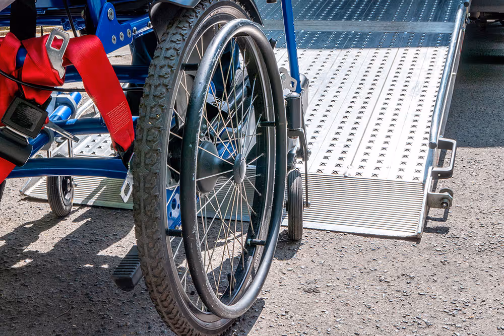 Close-up of a wheelchair wheel and red safety straps next to a metal wheelchair ramp on asphalt.