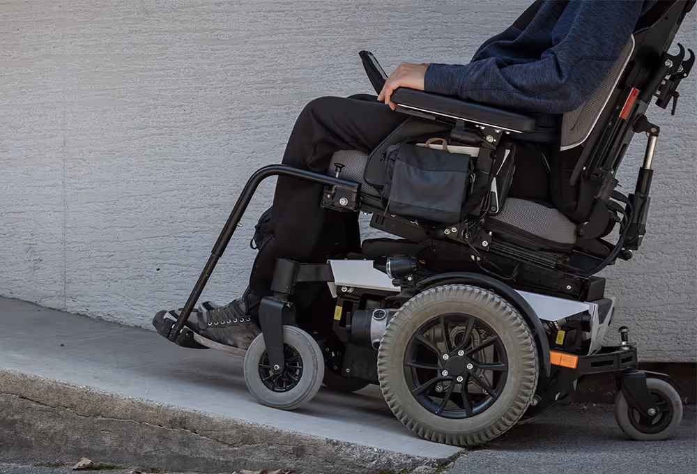 Person seated in a powered wheelchair on a concrete ramp next to a textured wall.