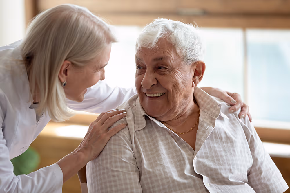 Smiling elderly man sitting as a woman gently places hands on his shoulders in supportive interaction.