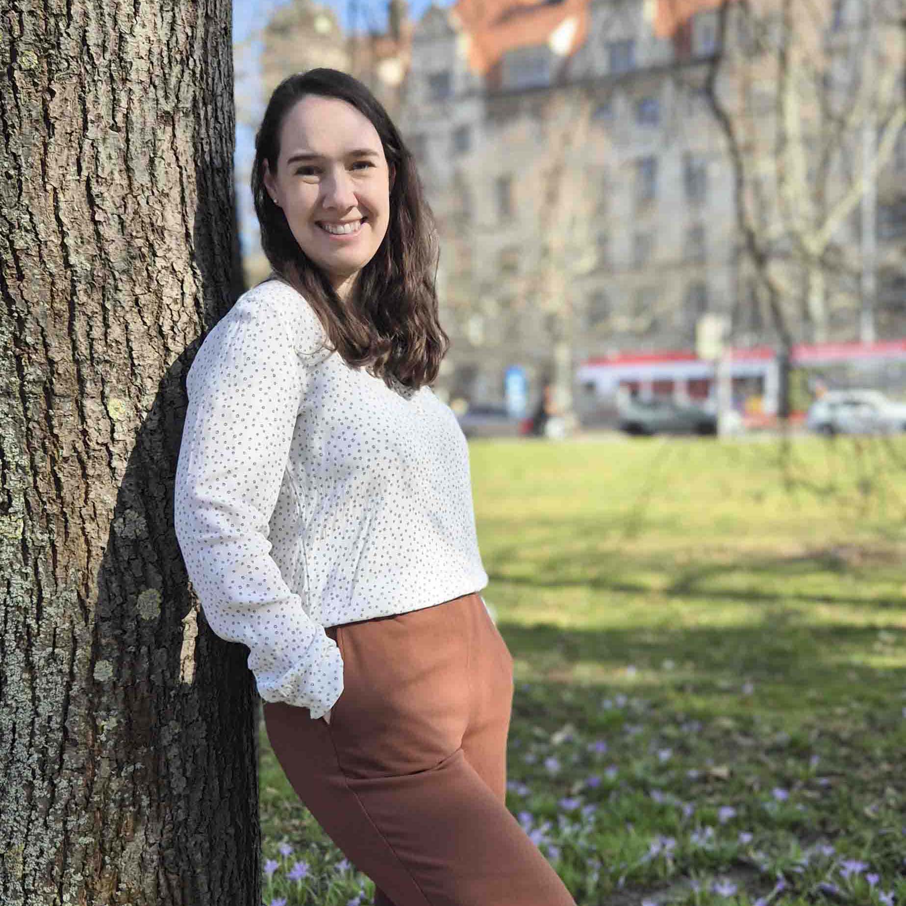 Frau lehnt lächelnd an einem Baumstamm in einem Park mit grüner Wiese und Blumen im Hintergrund.